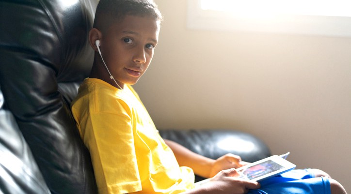 Foto um de menino, com blusa amarela e short azul, segurando um tablet e com um fone de ouvido