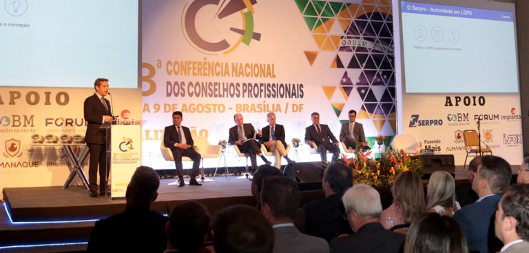 Presidente do Serpro, em pé, à esquerda, fala para o público do evento, assistido por cinco outros painelistas, sentados ao seu lado.
